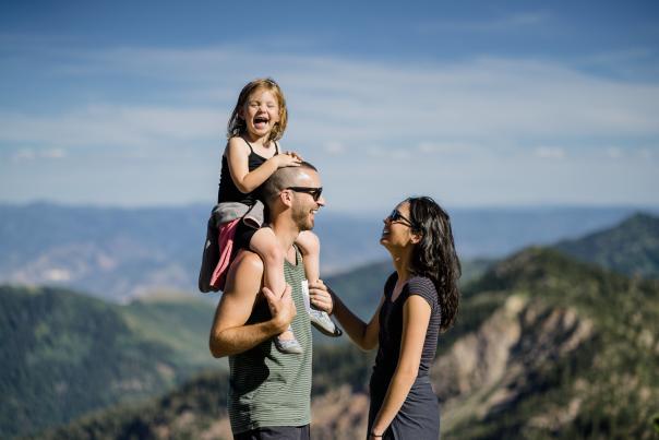 Man with happy child on his shoulders standing in front of a woman on a hiking trail mountaintop