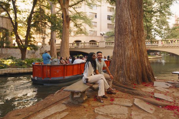 Couple sitting on bench at the River Walk in front of Go Rio RIver Cruise barge