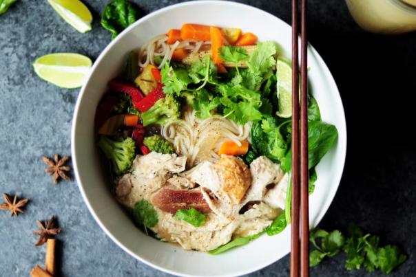 A white bowl with a noodle dish with chicken, broccoli, carrots, clinantro and peppers.