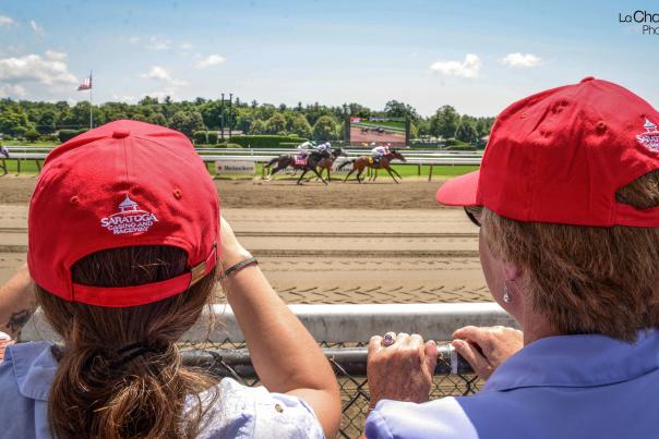 Two individuals in red Saratoga hats watching horses racing at Saratoga Race Course