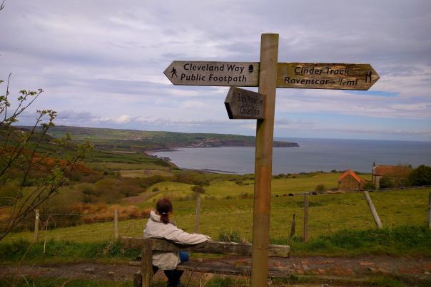 A blusery view from the cliffs looking over ravenscar, a sign points towards cleveland way public footpath, and a hiker takes in the view from a bench underneath it