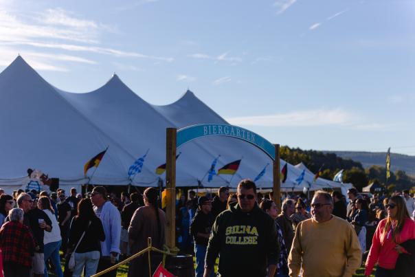 This image depicts a view of the crowd at the Glenville Oktoberfest, with a blue sky and forested hills in the background. A large biergarten tent can be seen, adorned by German and Bavarian flags.