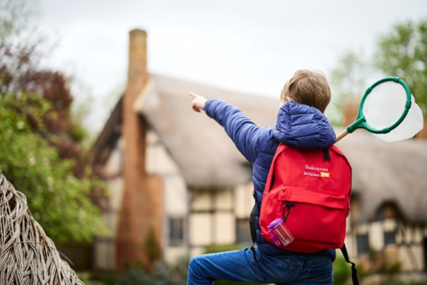 A boy with a red backpack on pointing towards Anne Hathaway's Cottage and holding a green net in the other hand
