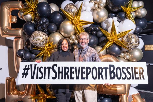 Man and woman standing in front of balloon display holding a #visitshreveportbossier sign