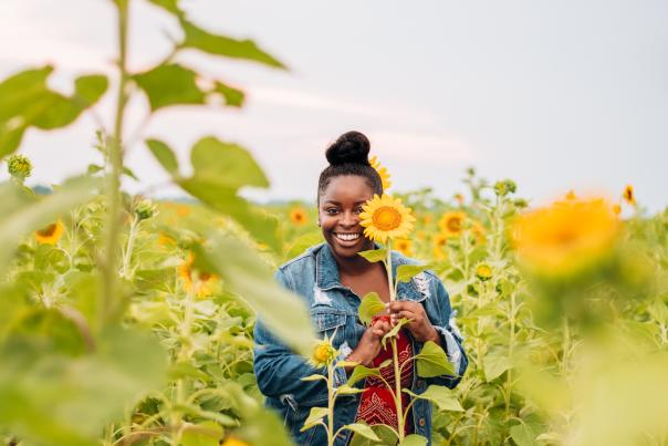 girl standing in the middle of sunflowers holding a sunflower over one eye and smiling