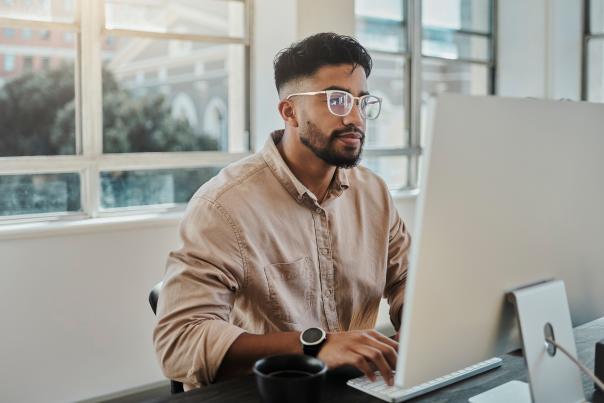 A young man with glasses and a beard sits designing at a desktop computer