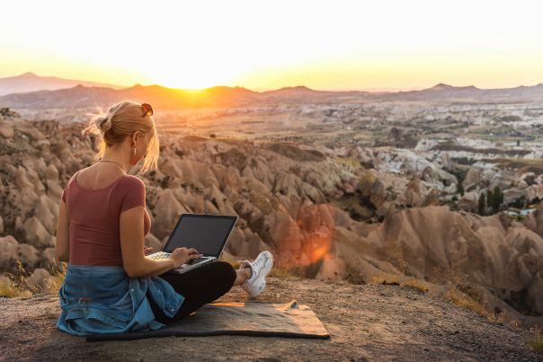A blong woman in a pink shirt with a denim jacket wrapped around her waste sites on top of a cliff and works on a laptop while the sun sets behind mountains in the distance