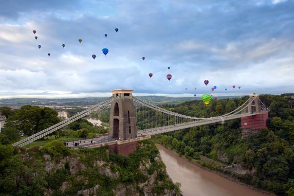 Hot air balloons hover over a bridge spanning a river.