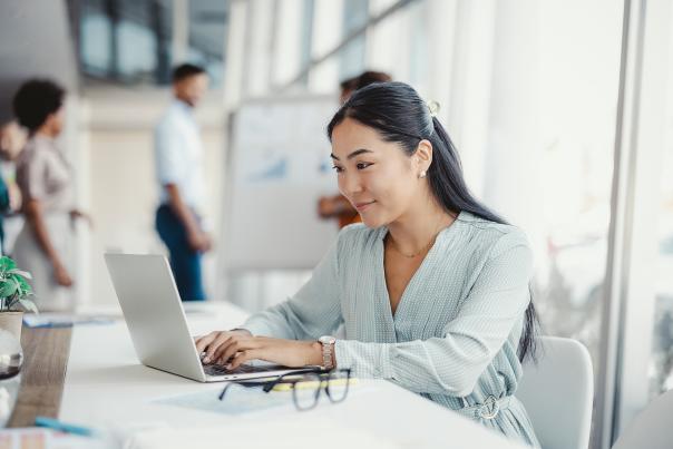 Businesswoman busy working on laptop computer at office with colleagues in the background.