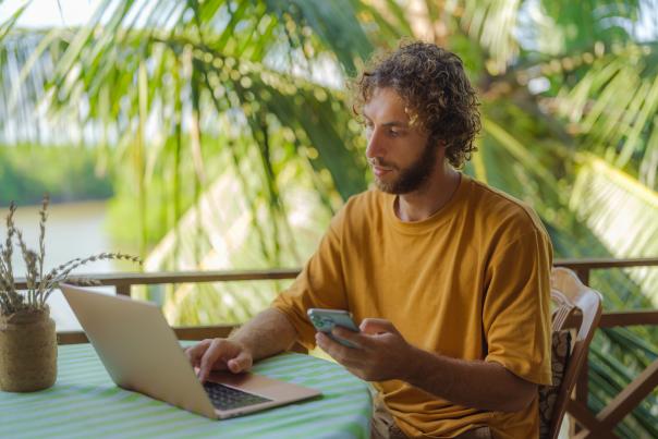 A man sits on a patio outdoors, surrounded by palm trees, while doing work.