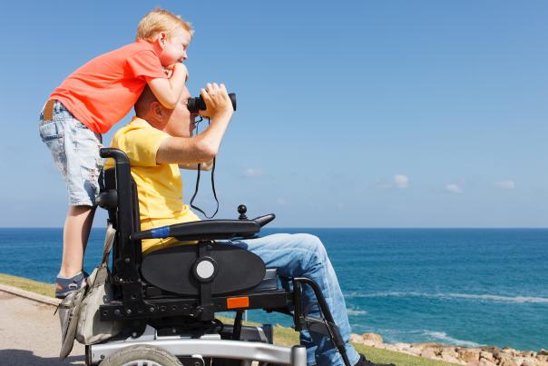 A young boy stands on the back of his father's wheelchair as he looks out with binoculars.