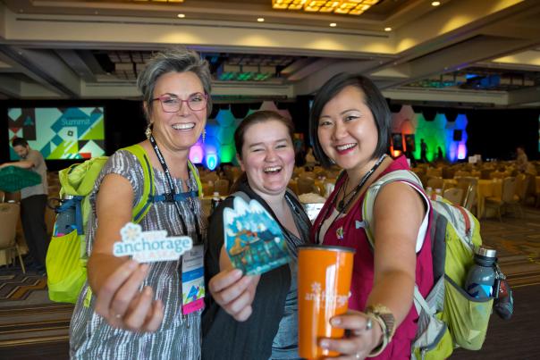 Three women smiling at the camera, holding up stickers and other swag items.