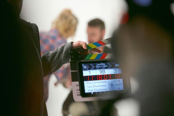 A movie clapperboard with two actors getting ready for a scene in the background.
