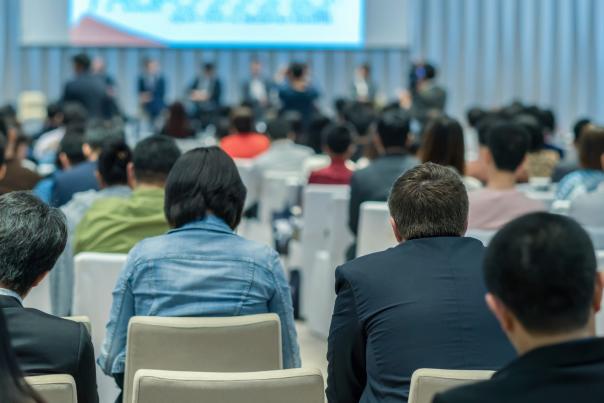 Conference attendees watch a panel discussion, with speakers on stage and participants seated in rows in a modern event hall.