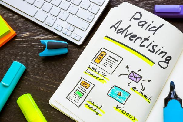 A keyboard and highlighters sit on a desk next to a notebook that has some drawings and the title, "Paid Advertising," on it.