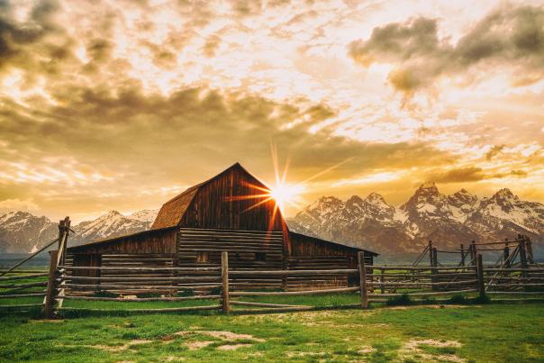 A barn in the forefront as the sun sets behind it and a mountain range.