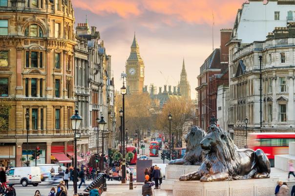 View of Big Ben and Westminster from Trafalgar Square in London at sunset, featuring historic architecture, lion statues, and red double-decker buses.