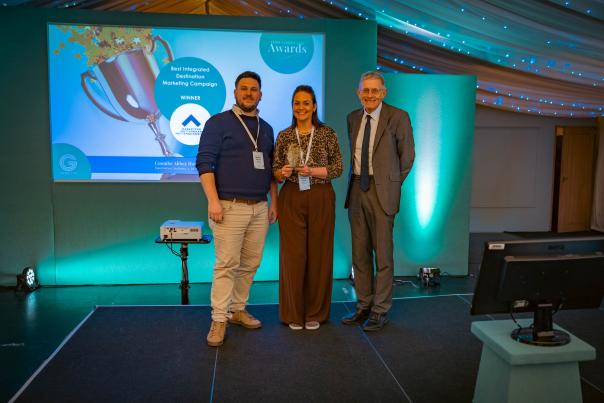 Three people on a stage holding an award at a marketing awards ceremony, with a winner announcement screen displayed behind them.