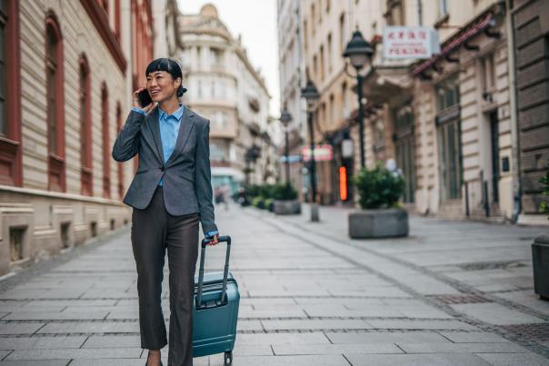 Business traveler with a rolling suitcase walking along a city street while talking on a mobile phone.
