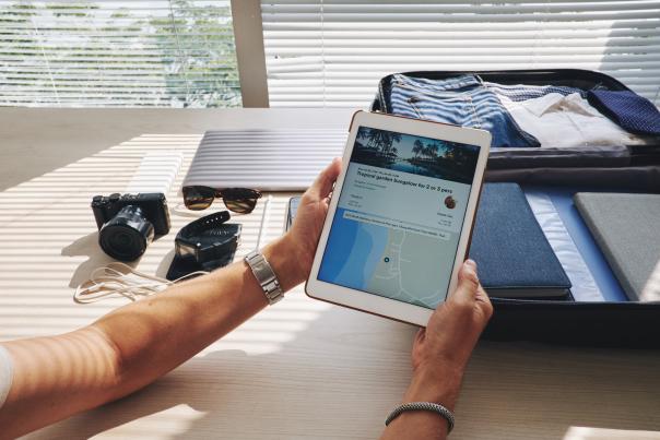 A person with a packed suitcase, camera, sunglasses and laptop laid out on a table browses accommodation listings on a tablet, representing local relevance in SEO for DMOs.