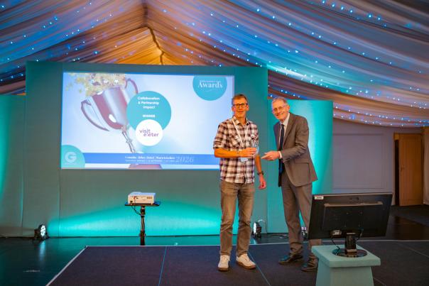 Award recipient accepts a trophy on stage at an awards ceremony, with a presentation screen showing a collaboration and partnership impact award.
