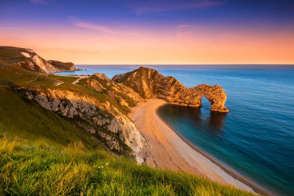 Coastal view of Durdle Door, a natural limestone arch on the Jurassic Coast in Dorset, England, with a sandy beach and calm blue sea under a vibrant sunset sky.