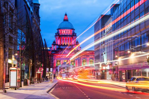 Belfast City Hall illuminated at dusk, with light trails from passing traffic along the surrounding city street.