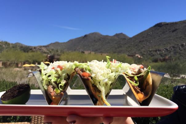 A person holds a plate of three tacos with Sonoran Desert foothills in the background.