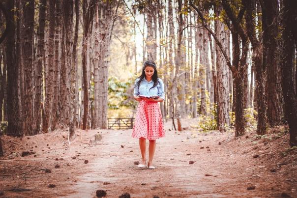 A woman walks down a path through the woods while reading a book.