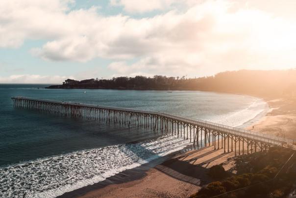 Long wooden pier stretches over blue ocean to a sandy beach at sunset, with warm sunlight, gentle waves and a California coast.