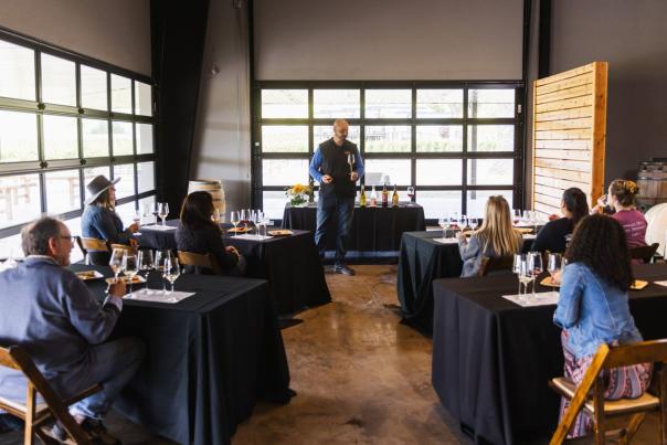 Man standing at the front of light filled room with a glass of wine and people sitting at tables in front of him