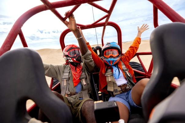Two people with helmets and goggles in a dune buggy with hands up