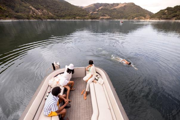 Three people in a pontoon boat watching one person swim alongside the boat in a lake