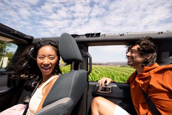 Two people smiling with open top car with wind in hair and fields in background