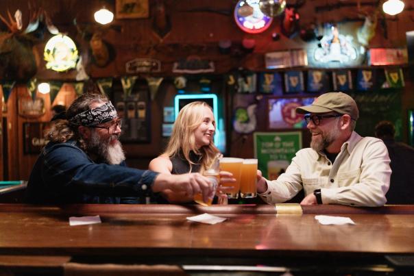 Three seated people cheersing will beer at a wooden bar with illuminated beer signs, flags and decor on the wall behind