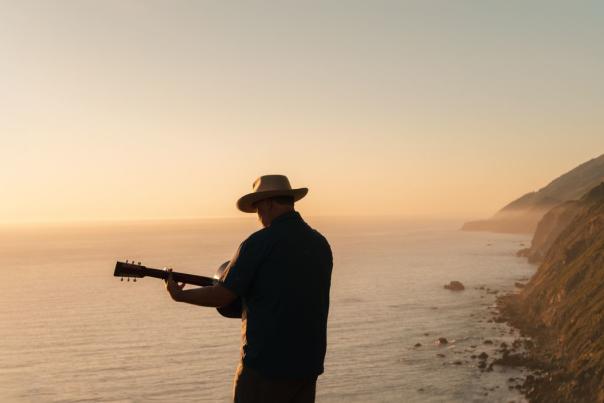 Man wearing a hat playing guitar with ocean and cliffside behind