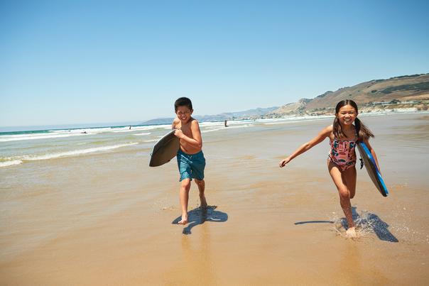 Two kids with boogie boards towards camera on the beach