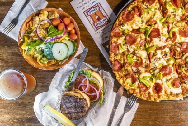 A wooden table featuring a large pepperoni pizza, a fresh garden salad, a burger with toppings, and a glass of beer.
