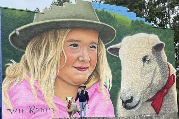 A mural of a girl with her pet lamb, with the actual girl and lamb posing in front in Winton