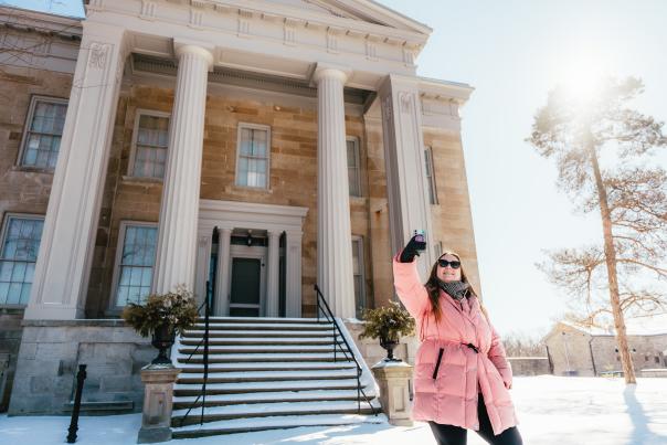 A woman wearing a pink winter coat and sunglasses smiles while taking a selfie in front of the mansion at Ruthven Park National Historic Site. Snow covers the ground and steps, and the bright winter sun shines through the clear blue sky.