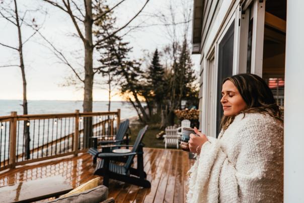 A person wrapped in a cozy white blanket stands in the doorway of a lakeside cottage, holding a warm mug and looking out over the water at sunset. A wooden deck with Adirondack chairs overlooks the shoreline, creating a peaceful late autumn atmosphere.