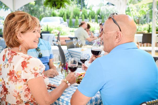 Two adults sit at an outdoor patio table under a white canopy, enjoying glasses of red wine and a meal while chatting with friends on a sunny day. Blue-and-white checkered tablecloths, flowers, and greenery in the background create a relaxed vineyard or garden dining atmosphere.