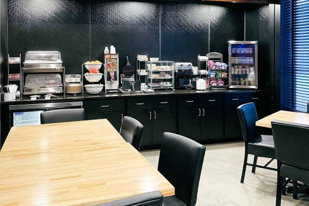 A square image of the cobblestone hotels breakfast bar. The counter is full of hot food, a waffle setup, cereal, a fridge with yogurts, milk juice, and a table in the front of the photo.
