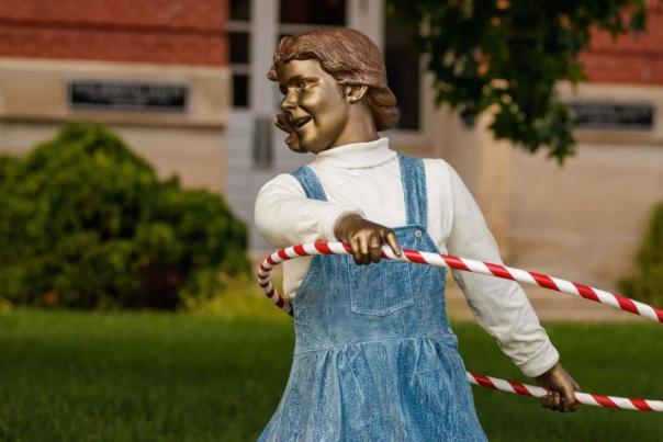 Life-sized bronze sculpture of a young girl twirling a red-and-white striped hula hoop, set on a grassy lawn in front of a brick building, capturing a playful moment in motion.
