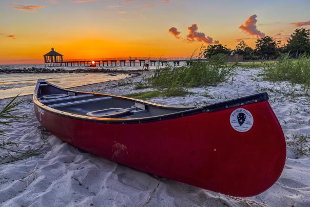 A red canoe rests on the white sand beach of Fontainebleau State Park at sunset with bushes and pier behind.