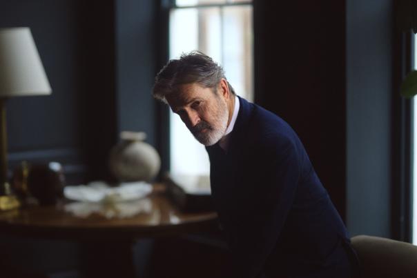 A portrait photo of Rupert Everett wearing a dark blue jacket, in a blue walled room