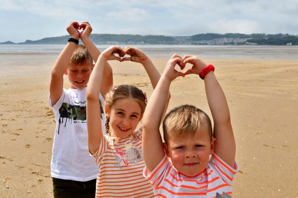 Three children on Swansea beach holding their arms in the air making a heart shape with their hands.