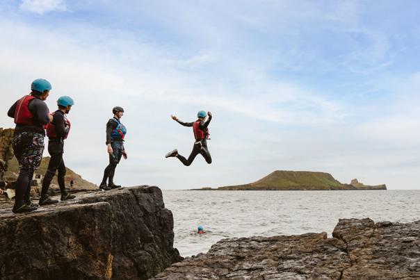 A person jumping into the sea wearing coasteering equipment with Worm's Head in the background.