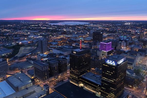 Syracuse city skyline at dusk with purple sky in the background, AXA towers and Barclay Damon building in the foreground, lit up purple.  City lights illuminate the photograph