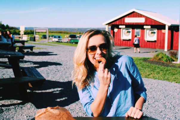 Young woman sitting at picnic table in front of red country store smiling while eating a cider donut